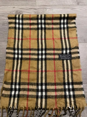 Klassisk rutig halsduk från Burberry - Säljer en ikonisk halsduk från Burberrys i beige, svart, vitt och rött rutmönster. Tillverkad i 100% lammull och har fransar i kanterna. Perfekt accessoar för att lyfta din outfit med klassisk brittisk stil. Hör av dig vid funderingar!:) snabbt köp = bättre pris! Fraktas samma dag som köp:)