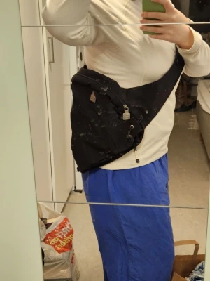 Björnborg axelväska  - Säljer en svart axelväska i tyg med flera fack och dragkedjor. Väskan har en bred rem som bärs över bröstet och praktiska detaljer som små spännen och snörning. Perfekt för dig som vill ha en enkel och funktionell väska till vardagen.