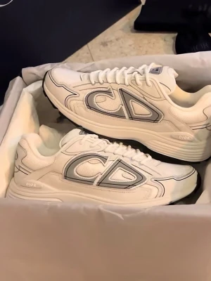 B30 vita sneakers - Säljer ett par sjukt snygga B30 sneakers i vitt med stora CD-loggor på sidan och diskreta grå detaljer. Skorna har en chunky sula, mesh och skinnpaneler samt klassisk snörning. Perfekta för dig som vill sticka ut med exklusiva och trendiga sneakers.