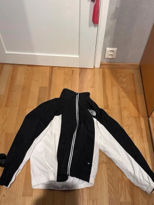 Svartvit vindjacka från The North Face - Snygg svartvit vindjacka från The North Face med hög krage och dragkedja framtill. Jackan har logga på bröstet och ryggen, samt elastiska muddar vid ärmslut och nederkant. Perfekt för blåsiga dagar och streetwear-stil. Orginal pris 1999 kr