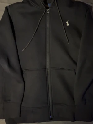 Svart hoodie från Polo Ralph Lauren - Klassisk svart hoodie från Polo Ralph Lauren med dragkedja framtill, justerbar huva med snören och fickor på magen. Ikoniska vita logobroderiet på bröstet. Perfekt för en chill och stilren look. Passar som small