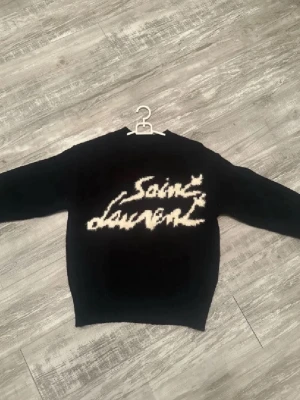 Saint Laurent tröja  - Säljer en svart stickad tröja från Saint Laurent med vit logotyp i graffiti-stil på bröstet. Tröjan har rund halsringning, långa ärmar och ribbade muddar. Perfekt för dig som vill ha en trendig och exklusiv look.