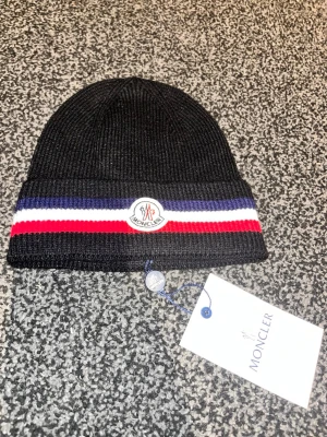 Svart Moncler mössa med ränder - Snygg svart ribbstickad mössa från Moncler med vikbar kant. Mössan har klassiska Moncler-loggan framtill och coola ränder i blått, vitt och rött runt om. Perfekt accessoar för dig som vill ha en stilren och trendig look.