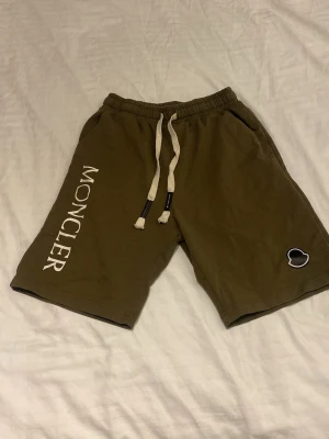 Olivgröna Moncler shorts med snörning - Snygga olivgröna shorts från Moncler med vit logga längs benet och snörning i midjan. Shortsen har två sidofickor och en liten Moncler-detalj på ena benet. Perfekta för sommaren och riktigt sköna att bära.