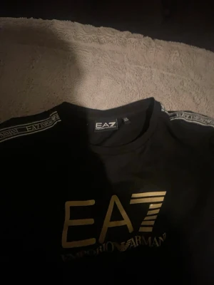 Svart EA7 Emporio Armani t-shirt - Snygg svart t-shirt från EA7 Emporio Armani med guldfärgat tryck framtill och logga på bröstet. T-shirten har korta ärmar och band med EA7-logga längs axlarna. Perfekt för dig som gillar stilrena och exklusiva plagg.