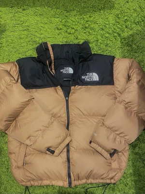 The North Face 700 - Säljer en beige och svart pufferjacka från The North Face. Jackan har hög krage, dragkedja framtill och logga på bröstet. Perfekt för kalla dagar med sin quiltade design och klassiska look.