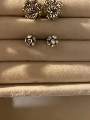 Mixade örhängen med pärlor och stenar - Säljer ett set med tre par örhängen: ett par stora silverfärgade studs med klara stenar, ett par små runda studs med multifärgade stenar och ett par tunna silverfärgade hoops med vita pärlor. Två par ringar en silver färgad med en snöflinga på en en helt vanlig guldringar.Perfekt för dig som gillar att variera din stil med både gnistrande och klassiska detaljer.Allt för 70kr,Ett par örhängen för 25 och en ring för 15kr