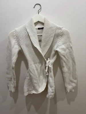 Vit omlottkofta  - Säljer en vit stickad omlottkofta med bred sjalkrage och knytband i midjan. Koftan har ribbstickade detaljer upptill och långa ärmar med muddar. Perfekt för lager på lager och en clean look.