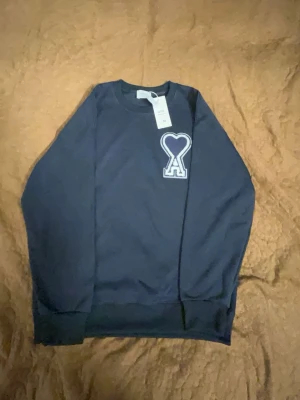 Mörkblå sweatshirt från Ami Paris - Mörkblå sweatshirt från Ami Paris med klassisk rund halsringning och ribbade muddar. På bröstet finns Ami Paris ikoniska hjärta-A-logga i vitt. Tillverkad i mjukt bomullsmaterial som känns skönt mot huden. Perfekt för en clean och stilren look.