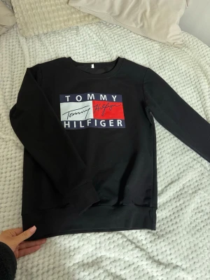 Svart sweatshirt Tommy Hilfiger - Svart sweatshirt från Tommy Hilfiger med stort tryck på bröstet i vitt, rött och marinblått. Klassisk rund halsringning och långa ärmar. Perfekt för en avslappnad och stilren look.