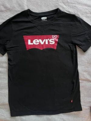 Svart Levi's t-shirt med logga - Klassisk svart t-shirt från Levi's med den ikoniska röda loggan tryckt på bröstet. T-shirten har rund halsringning och korta ärmar. Perfekt basplagg för en avslappnad och stilren look.