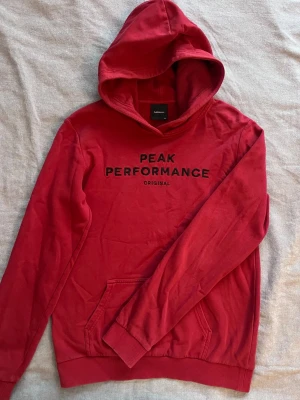 Röd hoodie från Peak Performance - Snygg röd hoodie från Peak Performance med svart text på bröstet. Klassisk huva och känguruficka framtill. Tillverkad i mjukt bomullsmaterial och har ribbade muddar vid ärmslut och nederkant. Perfekt för en avslappnad och sportig stil.
