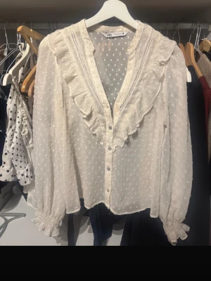 Vit volangblus med prickar från Zara - Supersöt vit blus från Zara med volangdetaljer längs v-ringningen och genomskinligt tyg med diskreta prickar. Blusen har långa ärmar med resår vid ärmslut och knäppning framtill. Perfekt för en trendig och romantisk look. Men en knapp har ramlat av