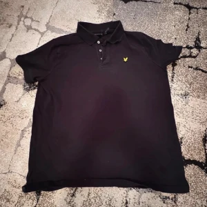 Svart pikétröja från Lyle & Scott - Klassisk svart pikétröja från Lyle & Scott med kort ärm och gul logga på bröstet. Perfekt för en stilren och avslappnad look. Tröjan har krage och knappar framtill.