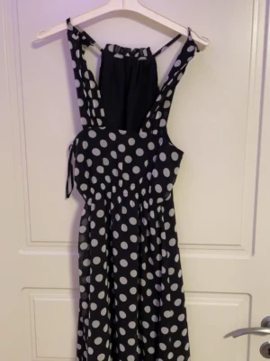 Svart klänning med vita prickar - Snygg svart klänning med stora vita prickar och smala axelband. Klänningen har en markerad midja, omlott-detalj vid axelbanden och ett flowy fall. Perfekt för dig som gillar retrovibbar och lekfull stil.