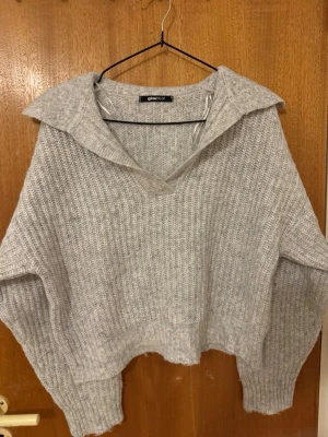 Grå stickad tröja från Gina Tricot - Mysig grå stickad tröja från Gina Tricot med bred krage och v-ringning. Tröjan har en avslappnad passform, ribbstickat material och långa ärmar. Perfekt för lager på lager och en chill look.