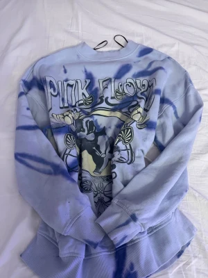 Ljuslila Pink Floyd sweatshirt - Säljer en ljuslila och blå tie-dye sweatshirt från DIVIDED med Pink Floyd-tryck framtill. Tröjan har rund halsringning, långa ärmar och ribbade muddar. Perfekt för dig som gillar bandtröjor och vill sticka ut med en unik färgkombination.