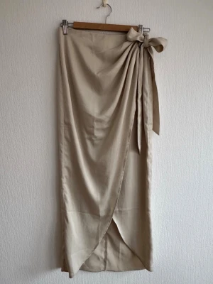 Beige omlottkjol med knytband - Stilren beige omlottkjol med knytband i midjan och lätt drapering framtill. 