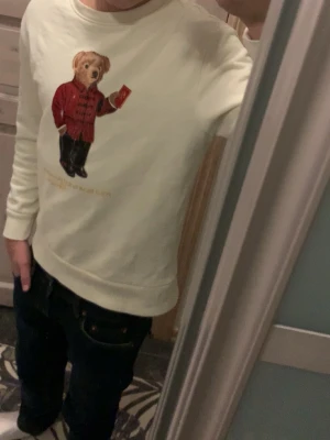 Vit sweatshirt med björntryck från H&M - Vit sweatshirt från H&M med ett stort tryck av en björn i röd jacka på framsidan, trycket har dock börjat lossna lite lite smått men inget som syns förens man verkligen kolla nära. Tröjan har rund halsringning och långa ärmar. Perfekt för dig som gillar lekfulla och trendiga plagg med en twist.