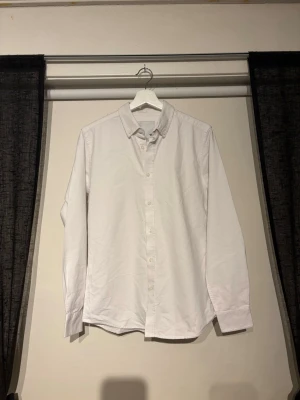 Klassisk vit skjorta med knappar - En stilren vit skjorta med lång ärm och knappar framtill. Skjortan har en klassisk krage. Enkel och tidlös design som alltid funkar.