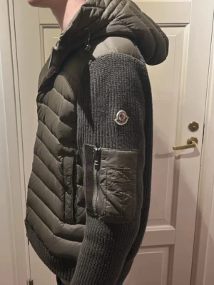 Grå Cardigan från Moncler med huva - Snygg grå teddyjacka från Moncler med stickade ärmar och vadderad kropp. Jackan har huva, dragkedja framtill och en praktisk ficka med dragkedja på ärmen. Moncler-logga på armen ger en exklusiv touch. Perfekt för kyliga dagar.
