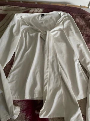 Vit omlottblus med knytband - Stilren vit omlottblus med långa ärmar och breda knytband framtill. Blusen har en djup v-ringning och är tillverkad i ett mjukt, lätt material som ger en avslappnad men chic look. Perfekt att styla med jeans eller kjol för en trendig outfit.