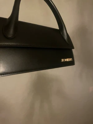 Jacquemus väska   - Säljer en stilren svart handväska/ armväska  från Jacquemus i slätt skinn. Väskan har ett elegant handtag, guldiga metalldetaljer och en minimalistisk design med loggan framtill. Perfekt för dig som gillar trendiga accessoarer och vill ha något som sticker ut.