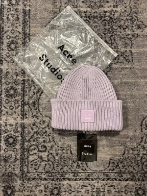 mössa Acne Studios - Säljer en ribbstickad mössa i ljuslila från Acne Studios med den ikoniska Face-loggan framtill. Mössan har bred uppvikt kant och är tillverkad i mjukt ull som håller dig varm och snygg. Perfekt accessoar för att lyfta din outfit.