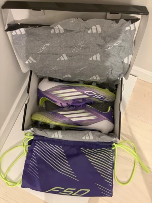 Adidas F50 lila fotbollsskor - Säljer ett par Adidas F50 fotbollsskor i lila, vitt och limegrönt. Skorna har snörning, klassisk låg profil och är tillverkade i syntetmaterial för lätt vikt och snabbhet på planen. Medföljer matchande påse med F50-tryck.