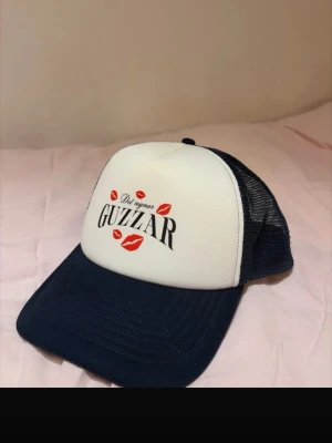 Papi santana keps  - Säljer en vit och marinblå truckerkeps med texten 'Det regnar GUZZAR' och röda läppavtryck framtill. Kepsen har mesh på sidorna och bak, samt böjd skärm. Perfekt för dig som vill sticka ut med en unik accessoar.