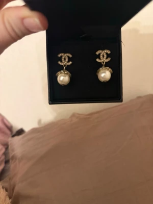 Örhängen med pärldroppe - Säljer ett par ikoniska örhängen i guld med glittrande stenar i logotypen och en stor vit pärldroppe. Perfekta för dig som vill ha något klassiskt. 