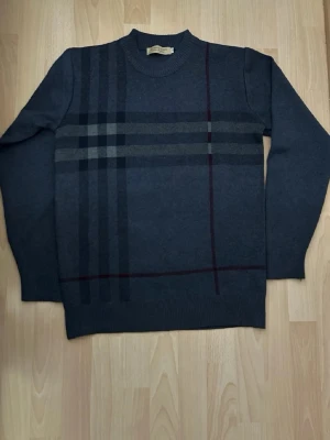 Mörkgrå stickad tröja från Burberry - Snygg mörkgrå stickad tröja från Burberry med klassiskt rutmönster i svart, grått och vinrött. Tröjan har rund halsringning och ribbade muddar vid ärmslut och nederkant. Perfekt för dig som gillar stilrena och tidlösa plagg.