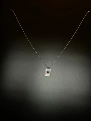 Halsband med hjärter ess-berlock - Snyggt halsband med silverfärgad kedja och en rektangulär berlock i form av ett spelkort, hjärter ess. Berlocken har en röd hjärtsymbol. Perfekt accessoar för dig som gillar unika smycken med korttema.