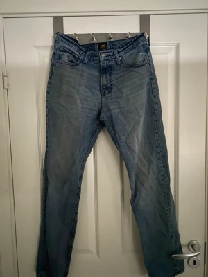 Blå  jeans från Lee - Klassiska blå jeans från Lee med rak passform och fyra fickor. Jeansen har en ljus tvätt och Lee-logga på bakfickan. Perfekta för en avslappnad och stilren look. Tillverkade i slitstarkt denimtyg av bomull. Den är dock lite sliten längst ner på benen på byxorna därför är den bra skick och inte mycket bra skick.