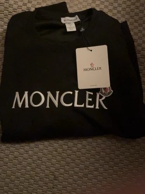 Svart sweatshirt från Moncler storlek xs - Stilren svart sweatshirt från Moncler med stort vitt Moncler-tryck på bröstet och rund halsringning. Tröjan har en klassisk passform och Moncler-logga på bröstet. Perfekt för dig som gillar exklusiva streetwear-plagg.