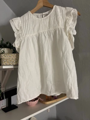 Vit blus med volangärmar - Söt vit blus med rund halsringning och volangprydda korta ärmar. Överdelen har fina veckdetaljer och spetskant, vilket ger en romantisk känsla. Tillverkad i ett lätt, luftigt tyg som känns skönt mot huden. Perfekt att styla med jeans eller kjol.