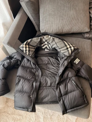 Svart dunjacka med huva från Burberry - Säljer en svart dunjacka från Burberry med klassiskt rutigt foder i huvan. Jackan har dragkedja och knappar framtill, två snedställda fickor med dragkedja och en patch med Burberry-logga på ärmen. Jackan går även enkelt att konvertera till väst. Perfekt för kalla vinterdagar, höst, vår och riktigt snygg streetstil.