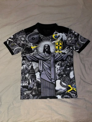 Brasil t-shirt med grafiskt tryck - Unik svart t-shirt från Nike med Brasil-logga och stort grafiskt tryck av Kristusstatyn och stadsmotiv. T-shirten har korta ärmar, inslag av gult och detaljerat mönster över hela framsidan. Storlek S men den är lite större i passformen så passar även M Perfekt för dig som gillar fotboll och streetstyle.