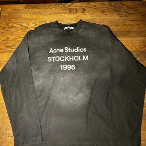Svart långärmad t-shirt Acne Studios (BAGGY) - Svart långärmad t-shirt från Acne Studios med vit text på bröstet där det står 'Acne Studios STOCKHOLM 1996'. Klassisk rund halsringning och relaxed fit. Perfekt för dig som gillar stilrena och tidlösa plagg.