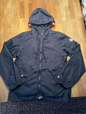 Marinblå vindjacka från Moncler - Snygg marinblå vindjacka från Moncler med huva och dragkedja framtill. Jackan har vita, röda och blå detaljer på huvan, två fickor med tryckknappar och Moncler-logga på ärmen. Tillverkad i lätt och tålig polyester, perfekt för blåsiga dagar.