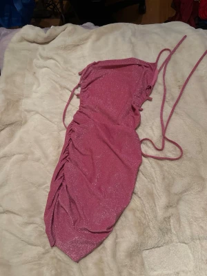 Glittrig rosa kort klänning - Säljer en glittrig rosa kort klänning med smala axelband och snörning i sidan. Klänningen har en åtsittande passform och rynkade detaljer längs ena sidan för en snygg effekt. Perfekt för dig som vill sticka ut med lite extra glitter.