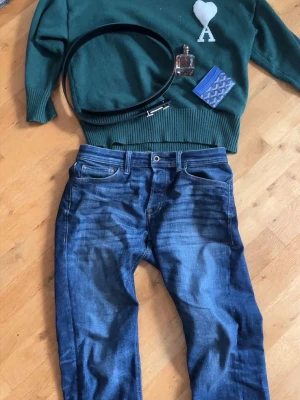 Grön stickad tröja från Ami Paris - Mörkgrön stickad tröja från Ami Paris med rund halsringning och ribbade muddar. På bröstet finns ett vitt hjärta med bokstaven 'A' under, vilket är Ami Paris ikoniska logga. Perfekt för dig som gillar stilrena och trendiga plagg.