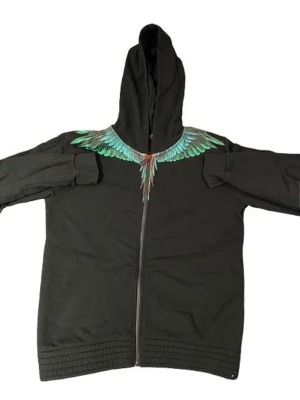 Svart hoodie från Marcelo Burlon - Säljer en svart hoodie från Marcelo Burlon med dragkedja och färgglatt vingmönster i grönt, blått och rött runt halsen. Hoodien har huva och ribbade muddar. Perfekt för dig som vill sticka ut med en unik design. Äkta givetsvis 