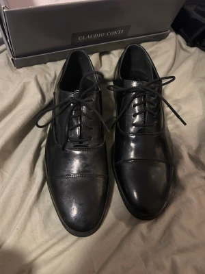 Svarta loafers från Claudio Conti - Stilrena svarta loafers från Claudio Conti i blankt skinn. Klassisk design med rund tå, snörning och diskreta sömdetaljer över tån. Perfekta för dig som vill ha en tidlös och elegant look till vardags eller mer uppklädda tillfällen. Endast använda ca 1-2 gånger.