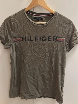 Olivgrön Hilfiger t-shirt XS - Snygg olivgrön t-shirt från Tommy Hilfiger med tryckt logga och texten 'HILFIGER NEW YORK' på bröstet. T-shirten har korta ärmar och klassisk rund hals. Tillverkad i mjuk bomull, perfekt för en avslappnad och trendig look.
