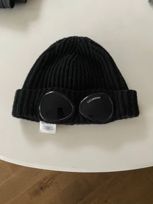 Svart mössa med goggles från C.P. Company - Säljer en svart ribbstickad mössa från C.P. Company  Mössan är i tjockt,mjukt material och har en uppvikt kant där glasögonen sitter fast. De är bara o fråga om fler bilder 