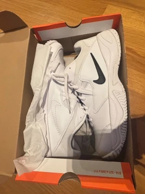 Vita Nike sneakers med svart swoosh - Säljer ett par fräscha vita Nike sneakers med svart swoosh på sidan. Skorna har klassisk snörning, rund tå och platt sula. Tillverkade i syntetmaterial och har en sportig, stilren look som passar perfekt till streetwear.