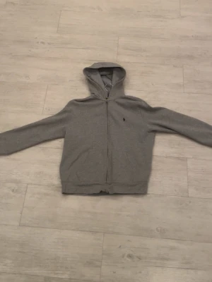 Grå zip hoodie från Polo Ralph Lauren - Klassisk grå hoodie med dragkedja från Polo Ralph Lauren. Tröjan har huva, långa ärmar och två fickor framtill. Liten broderad logga på bröstet. Tillverkad i mjukt bomullsmaterial som är skönt att ha på sig.