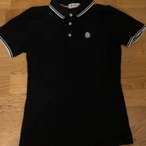 Svart Moncler pikétröja med krage - Svart pikétröja från Moncler med klassisk krage och vita ränder på krage och ärmslut. Tröjan har tre knappar framtill och Moncler-logga broderad på bröstet. Tillverkad i mjuk bomull, perfekt för en clean och stilren look.