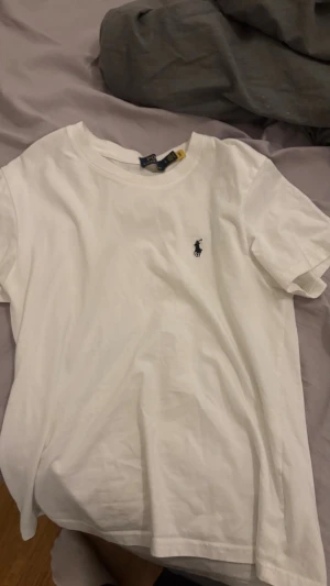 Vit t-shirt från Polo Ralph Lauren - Vit t-shirt från Polo Ralph Lauren i mjuk bomull. Klassisk rund hals och korta ärmar. På bröstet finns den ikoniska broderade loggan i blå. Enkel och stilren design som funkar till allt. Perfekt för dig som gillar clean och tidlös stil.
